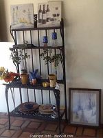Wide view of black metal stand with shelves holding various faux flowers, decorative bowls, plates, and standing canvas pictures on top, plus separate framed winter scene artwork leaning against wall.