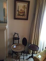 Four small side tables grouped near a wall under a framed painting, showing two wooden tables, one gold tray table, and black oval wooden table top leaning on wall