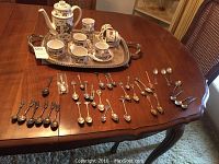 Full view of tea set including tea pot, six cups and saucers on tray, creamer and sugar bowl, with a large collection of assorted spoons spread on table in front