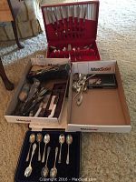 Overview of red velvet cutlery chest, fitted black case of spoons, two MaxSold trays with loose silver-plate flatware and boxed cake knife/fork.