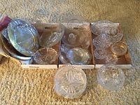 Lot showing six solid brass serving plates stacked, assorted crystal bowls and vintage glass ashtrays in cardboard boxes on carpeted floor.