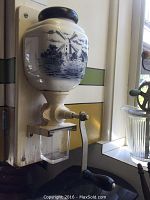 Photo of the Holland-themed wall-mounted coffee grinder with ceramic container decorated with a windmill scene, attached to a wall with visible crank handle.