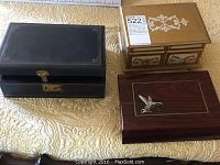 Three closed jewelry boxes: one black/blue, one gold with floral designs and two drawers, and one wooden with bird inlay.
