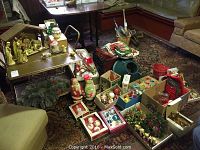 Wide view of multiple vintage Christmas decorations arranged on a patterned carpet including boxes of ornaments, light-up Santa and snowman, wreaths, and other decor near furniture.