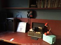 Wide shot including Philips reel to reel recorder with headphones, blue radio, orange desk lamp, papers, rotary phone and bookshelf above the shelf