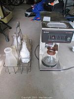 Industrial Bunn coffee maker with glass carafe next to metal carrier holding eight glass milk bottles with some residue inside.