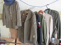 Wide view of several WWII US military uniforms on hangers, showing Army and Navy jackets with patches and insignias.