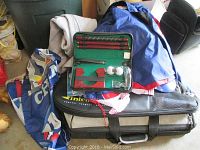Photo of mixed sports fan gear including a blue and red New York Giants jacket, a folded garment, part of a golf putting practice set in black carrying case and other fan apparel items.