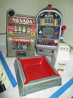 Front view of the metal Nevada slot machine, electronic Ultimate Casino slot machine, Harrah's ceramic bank, and Courvoisier cognac ashtray arranged on a surface.