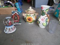 Overview photo of the lot showing the covered jar with sunflower design, Fitz and Floyd flower vase, two holiday snow globes (house-shaped and carousel Santa Claus), and clear measuring cup