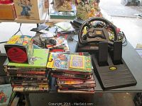 View of Mad Catz Dual Force racing wheel with pedal set and stacked game cases on a table showing various video games and accessories in the lot.