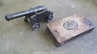 Wooden cigar case with logo and metal cannon model side by side on floor.