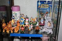 Photo showing an assortment of Disney figurines including Lion King characters (Simba, Mufasa, Timon, Pumbaa), Donald Duck, Pluto, Snow White figurines, and a pink Holly Hobbie thermos with floral umbrella design on a glass shelf.