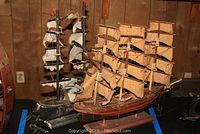 Photo shows two wooden model ships on display on a table with wood panel background. The ship on the right is labeled Fragata Espanola with multiple sails and rigging. The ship on the left is an older styled wood ship model with black base.
