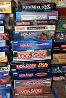 Stacked boxed board games showing titles Rummikub, Amend-OPOLY, multiple Monopoly editions including Pirates of the Caribbean, Washington D.C., Disney Pixar, Star Wars, Looney Tunes, Lord of the Rings, Americana, Deluxe, and .com Edition.