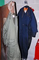 Three medium snowmobile suits hanging on hangers against a wall. Includes green one with fur-lined hood, blue one with orange lining, and black one with red collar lining.