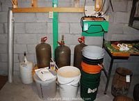 Photo of various buckets stacked and large glass carboys with valves, along with a measured pail containing white powder and a small can of additive on the floor.