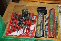 Close-up of various drill bits including spade and auger bits alongside some drill bit packaging.