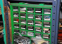 Green plastic storage caddy with many small transparent drawers labeled and filled with various metal hardware, nuts, washers, and spacers.