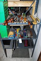Full view of three metal shelves with wooden-handled rasps/files on the top shelf, various tools and items on middle and bottom shelves.