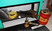 Photo showing the gasoline can, chimney brush with yellow rope, two wooden-handled saws, brass blowtorches, and machete arranged on a shelf and floor against a light wall.
