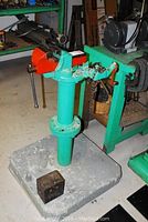 Overview of entire ridge pipe bender unit showing concrete base, teal stand, orange vise clamp, and mechanical parts.