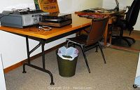 View of collapsible table with wooden top and black metal folding legs, brown metal-framed chair, black office chair, and metal waste can beneath the table.