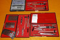Four incomplete drafting tool sets laid out with cases open, showing compasses, dividers, and metal stencils on a wooden table.