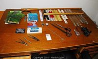 Overview of drafting tools and accessories arranged on wooden table, including rulers, compasses, shears, mechanical pencil leads, T-squares, and drafting stencils.