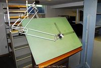 Side view of the drafting table showing the green angled drawing surface, articulated arm, and wooden base.