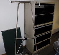 View of beige metal A-frame easel and green chalkboard panel leaning against it with vertical filing cabinet in background.