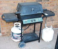 Front view of Broil-Mate BBQ grill with two burners, side shelves, two propane tanks attached below, showing wheels and stand.