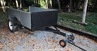 Overall view of steel frame trailer bed with plywood side panels and attached tow dolly on gravel surface