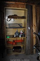 Full view of beige metal shelving unit containing rope, cage trap, funnels, clamps, ammo box, with wooden planks alongside.