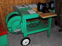 Full view of green metal cart with wheels. Jobmate bench grinder mounted on wooden board on top shelf. Bench vice attached to cart. Manuals laid out on top shelf. Large black extension cord wound on cart side.