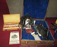 Wide shot of Black & Decker router with attached cord, placed inside wooden case lined with blue fabric, next to wooden box containing router bit set and instruction manual.