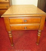 Front angle of wood end table showing top, two drawers with metal knobs, and turned legs on red carpet.