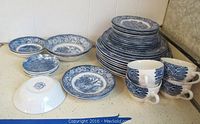 Various sizes of Liberty Blue china plates and bowls with blue and white pattern; some stacked. Four teacups with handles also included.