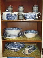 Cabinet shelf displaying Liberty Blue china: two covered pitchers, three drinking glasses, bowls, and plates.