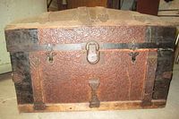 Front view of wooden trunk showing embossed floral design on front and central metal lock, missing side handles