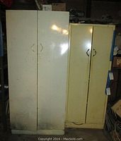 Two metal workshop cabinets shown side by side in a basement setting, a taller white cabinet on the left and a shorter yellow cabinet on the right, showing signs of wear and dirt on surfaces.