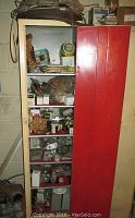 Tall red metal cabinet with multiple shelves holding various electrical components including bulbs, wiring devices, and boxes of parts