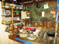 Photo of wooden workbench with a bench grinder mounted, assorted hand tools, scissors, pliers, wire spools, and hardware items on shelves and pegboard in workshop corner.