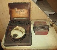 Antique Jewell Standard volt meter in open wooden box with probe wires inside, paired with a red metal train transformer with attached cord.