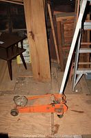 Side view of orange floor car jack with white handle on plywood floor with wooden furniture and ladder in background.