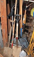Three shovels with red paint on handles leaning against a wall with a metal rake behind. Shows condition and types of garden tools present.
