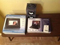 Photo showing the boxed VisionQuest 7-inch digital photo frame, boxed Lloyd's 9-inch portable DVD player, and two speakers (Vibe kids speaker and unknown speaker) on a wooden surface.