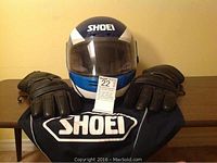 Front view of the vintage Shoei helmet with clear visor down, flanked by leather riding gloves, and Shoei helmet cover spread below.