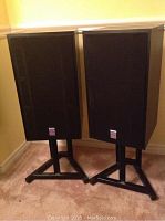 Full view of two black Cerwin-Vega speakers on black stands placed side by side on carpeted floor in a corner with beige walls.