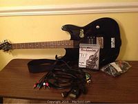 Full view of black First Act electric guitar with strap, Rocksmith PS3 game in case, and universal RCA cables with selector switch on table.